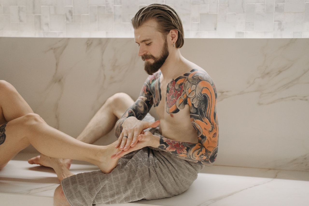 our-mission-01 A tattooed man receives a foot massage indoors, showcasing vibrant body art.